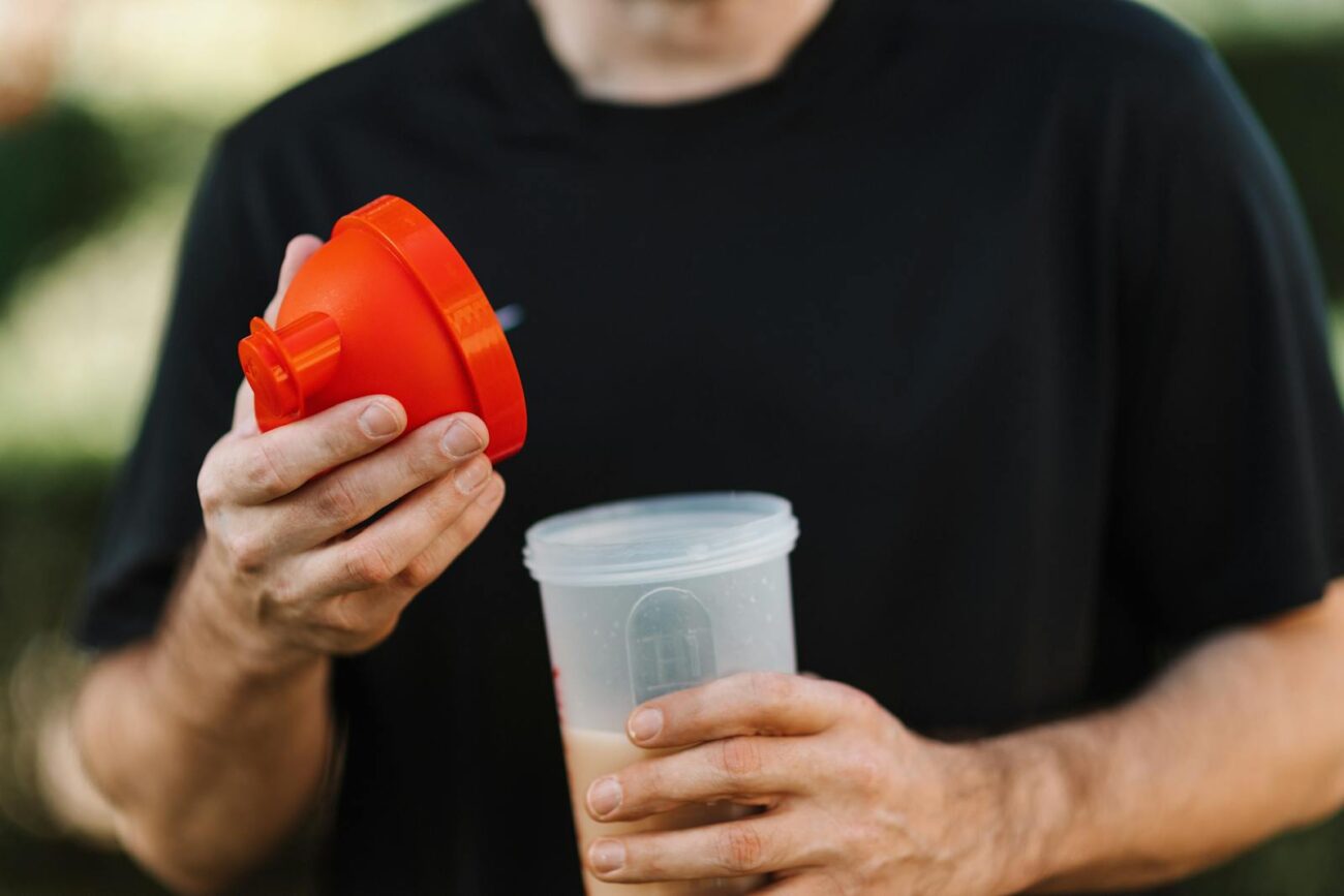 A man in a black shirt holds a protein shake tumbler with an orange lid, outdoors.