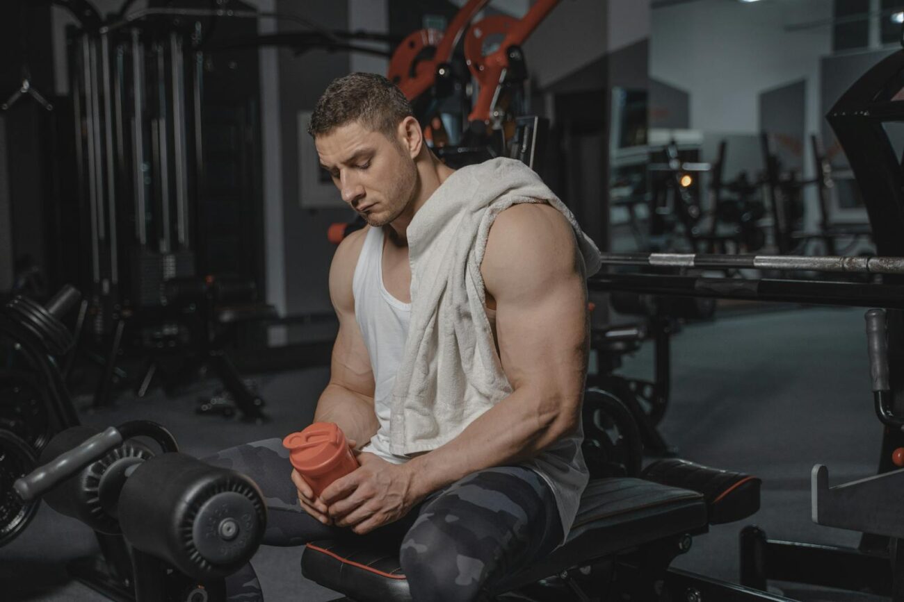 Muscular man sits in gym holding protein shaker after a workout, towel on shoulder.
