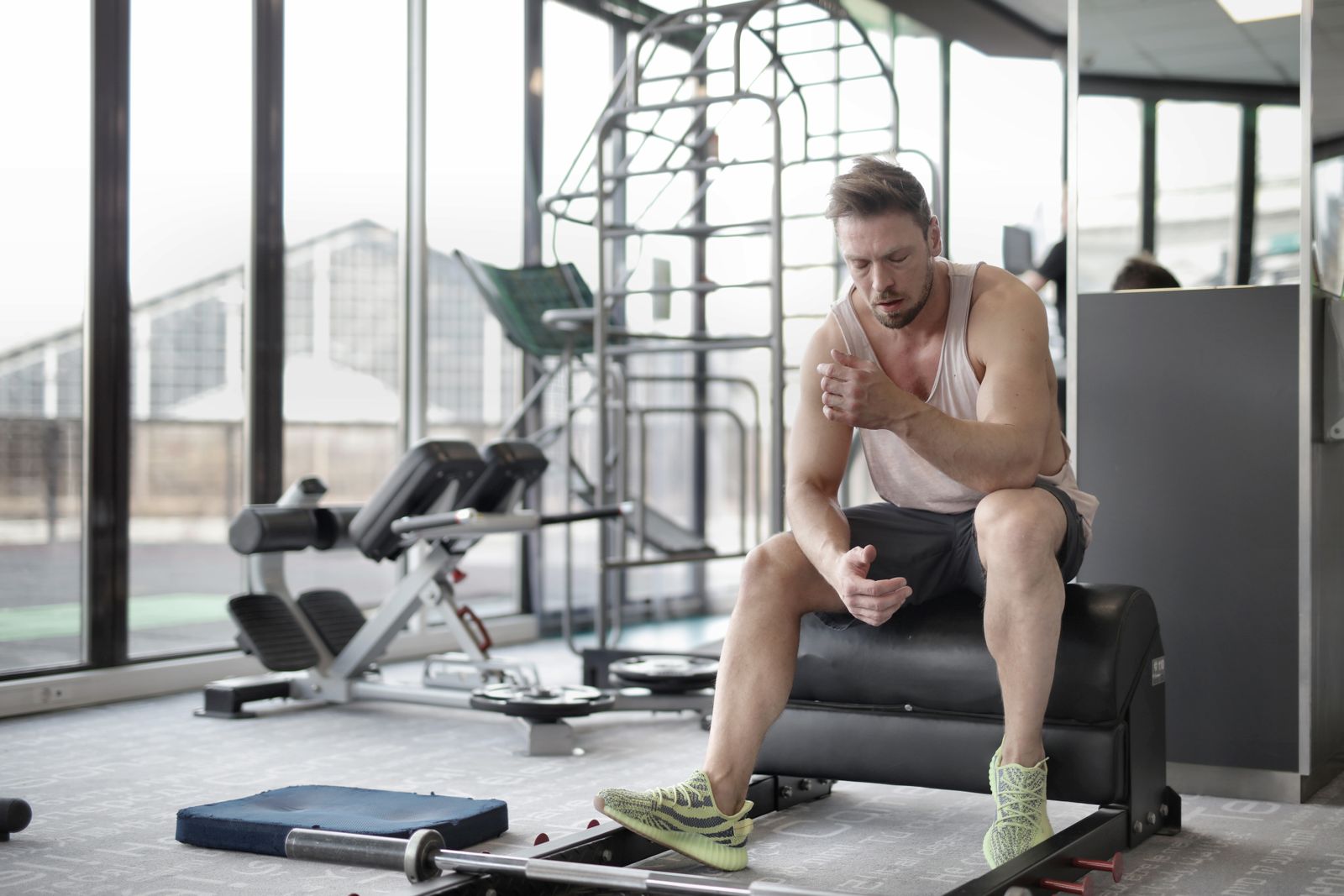 A man sitting on a gym bench catching his breath during a rest break between sets, with a barbell on the floor