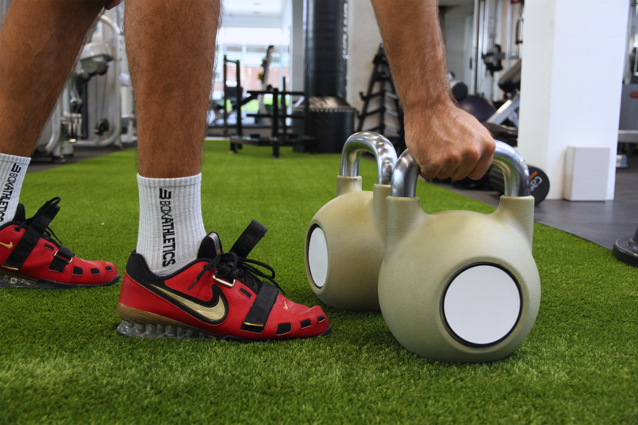Athlete reaching down to pick up a light beige competition kettlebell on green turf in a gym