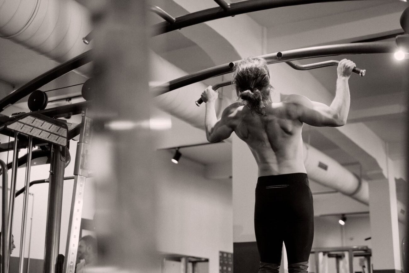 Back view of a shirtless athlete performing wide-grip pull-ups on an overhead pull-up bar frame in a gym