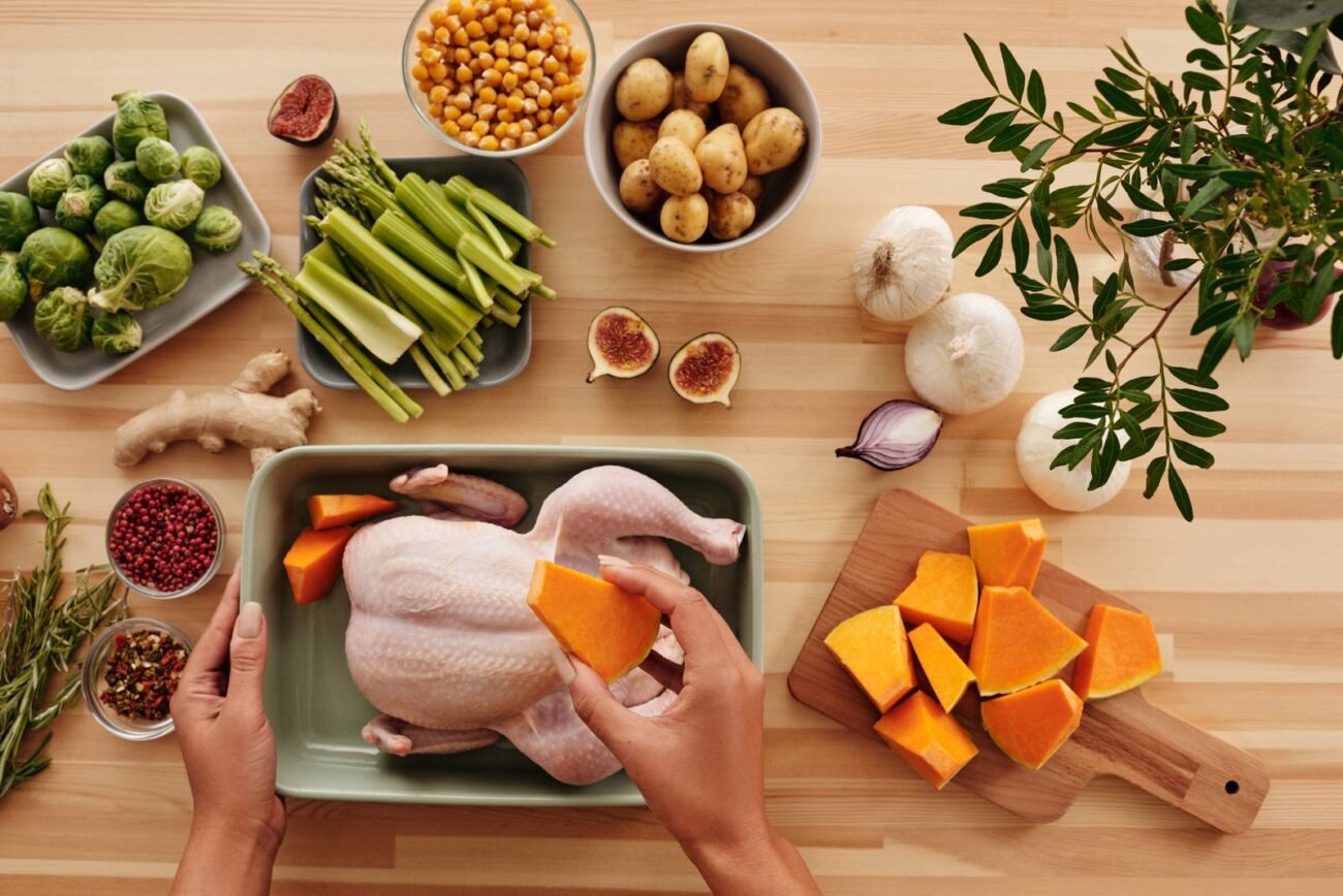 Top-down view of hands arranging raw chicken and chopped squash in a baking dish surrounded by fresh vegetables