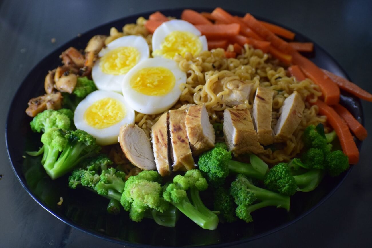 Plate filled with noodles topped with sliced grilled chicken breast, hard-boiled eggs, broccoli, carrots, and mushrooms