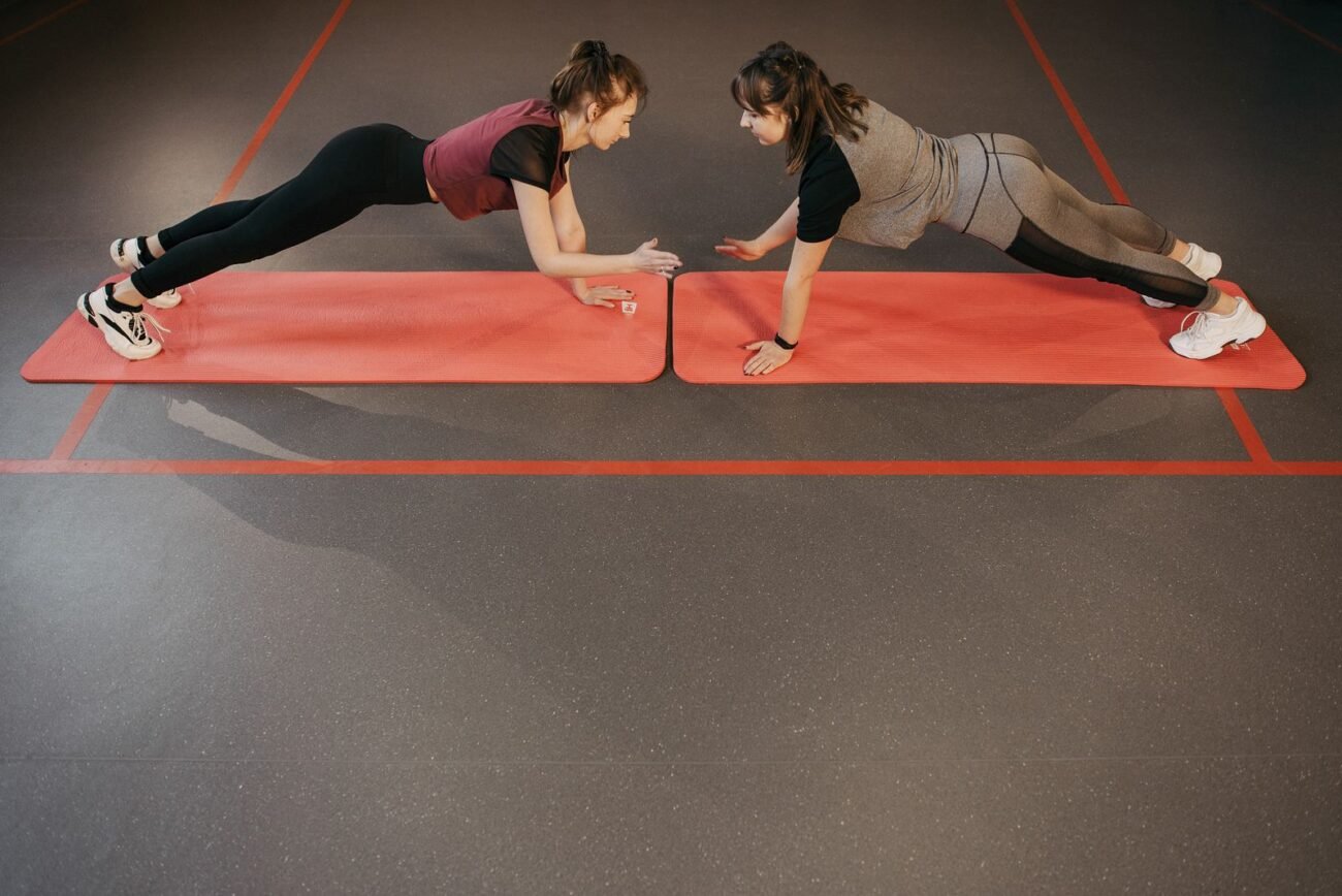 Two women facing each other while holding high planks on red non-slip fitness mats during a joint workout